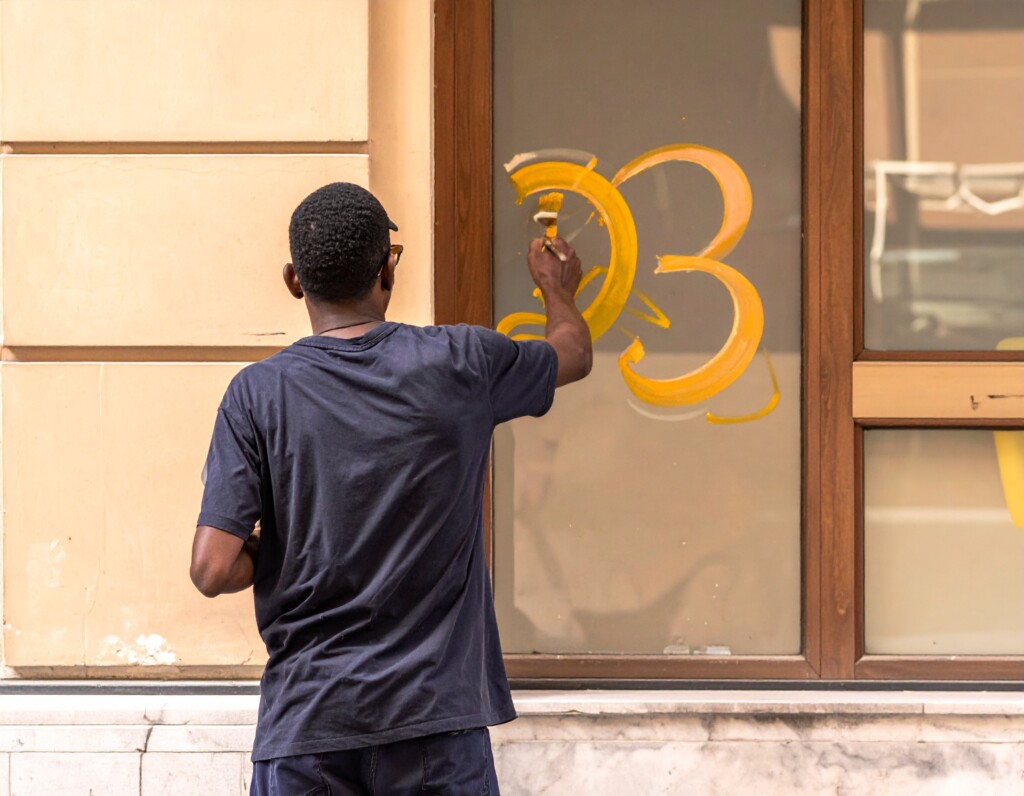 Un peintre en lettres de dos entrain de réaliser un lettrage sur une vitrine dans la rue